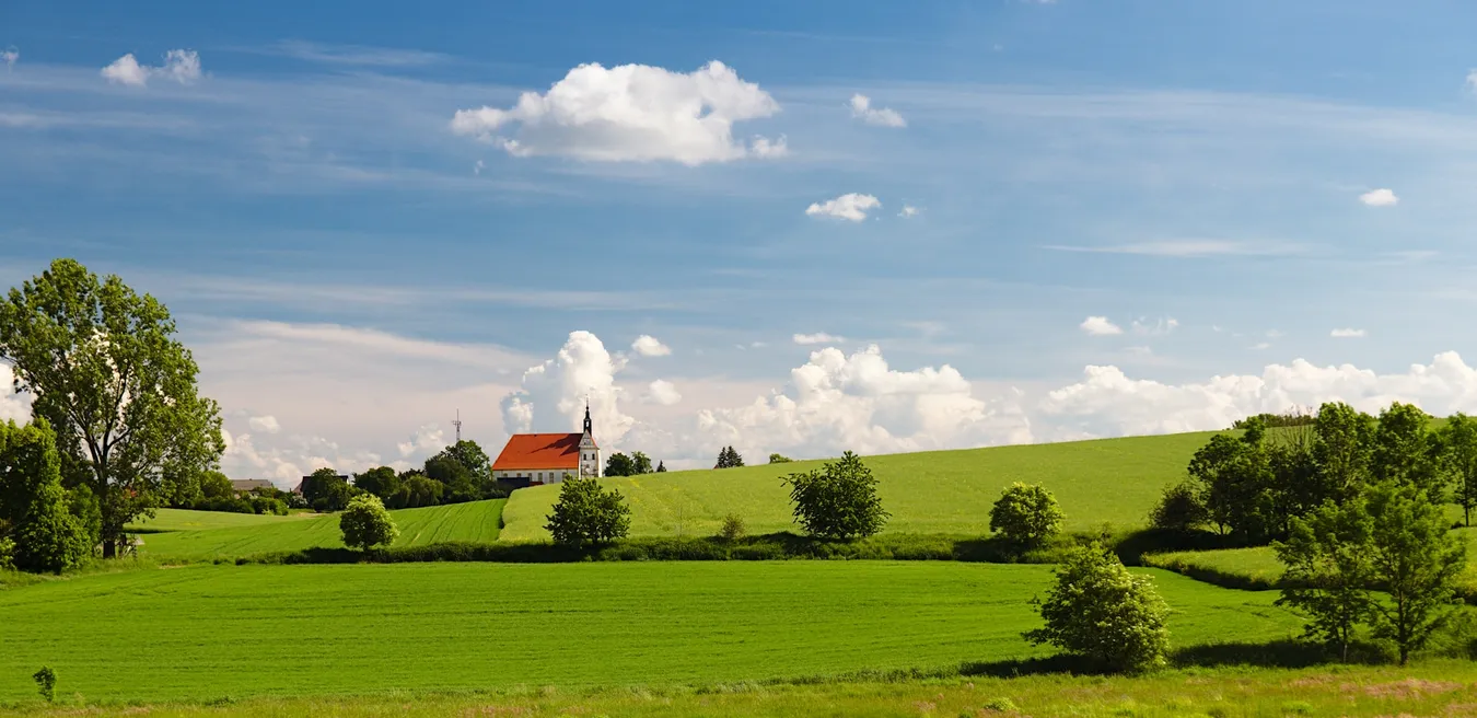 Panoramablick auf eine weite, sattgrüne Wiese mit vereinzelten Bäumen; im Hintergrund steht eine kleine, weiß getünchte Kirche mit rotem Dach auf einem sanften Hügel, darüber ein strahlend blauer Himmel mit weißen, flauschigen Wolken.